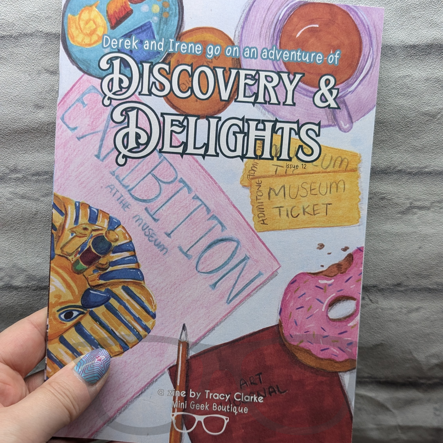 Zine titled 'Discovery & Delights' held by a hand with a Gray background. The zine features a much longer story, an article, activity pages and a special comic by This Goblin Makes