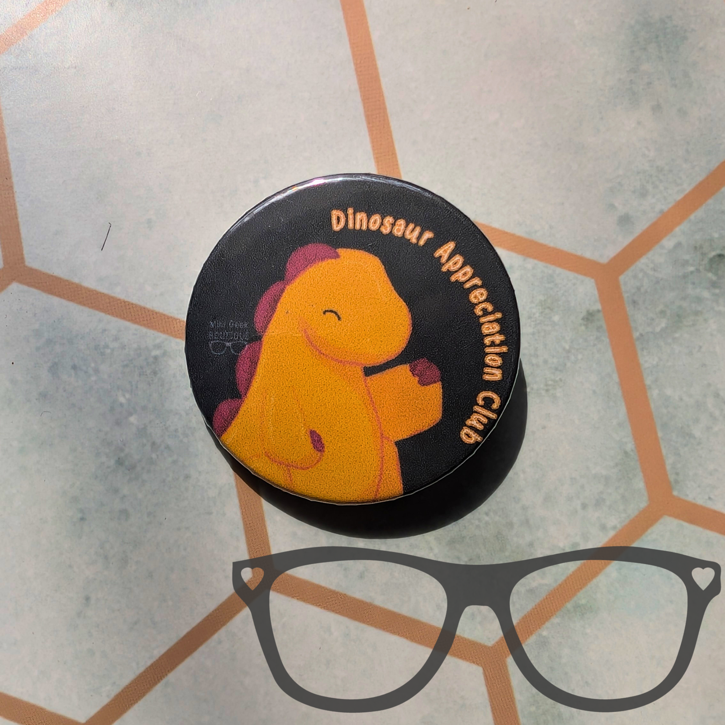 A round black pinback button or sticker featuring a cartoon yellow dinosaur with purple spikes and the text 'Dinosaur Appreciation Club,' placed on a light grey patterned surface. A grey glasses outline graphic is overlaid at the bottom.