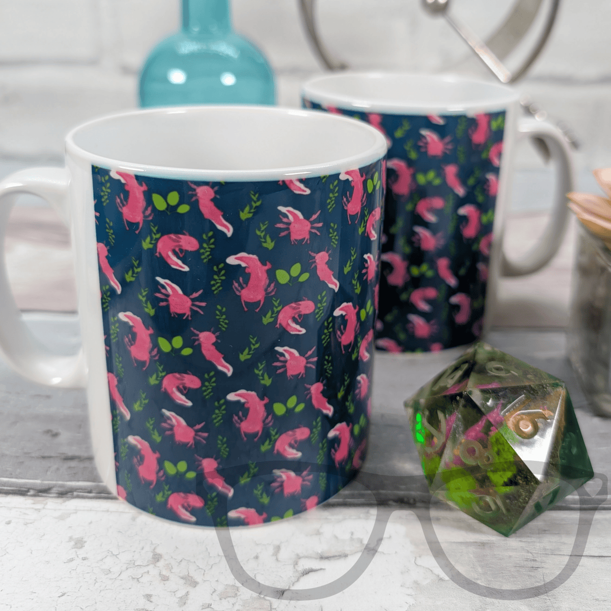 Two mugs with pink and green axolotl designs on a surface with a dice in the foreground.