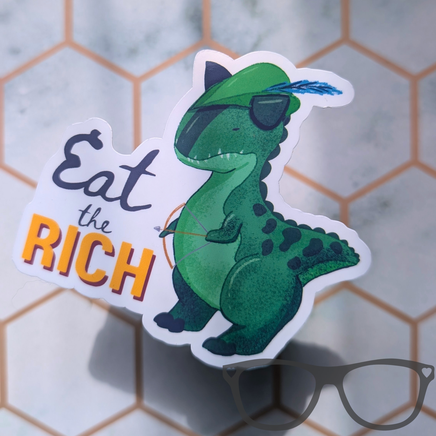 A die-cut sticker featuring a green illustrated Carnotaurus a Robin Hood and a blue feather in its green hat, next to the text 'Eat the RICH' in grey and orange lettering.