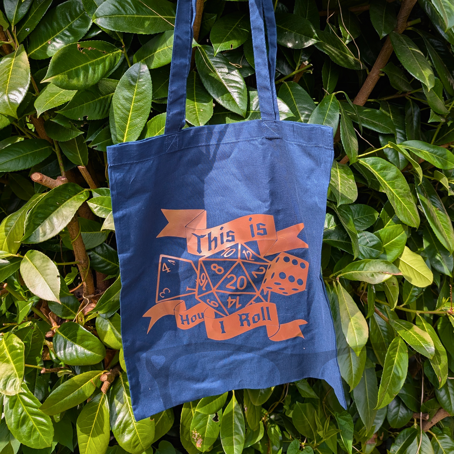 Navy blue tote bag with rose gold vinyl design reading "This is how I roll," featuring various gaming dice, hanging from a lush green bush. A stylish and playful accessory perfect for tabletop gaming fans on the go.