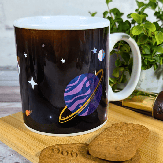 Mug with space-themed design on a wooden surface with cookies and a plant in the background