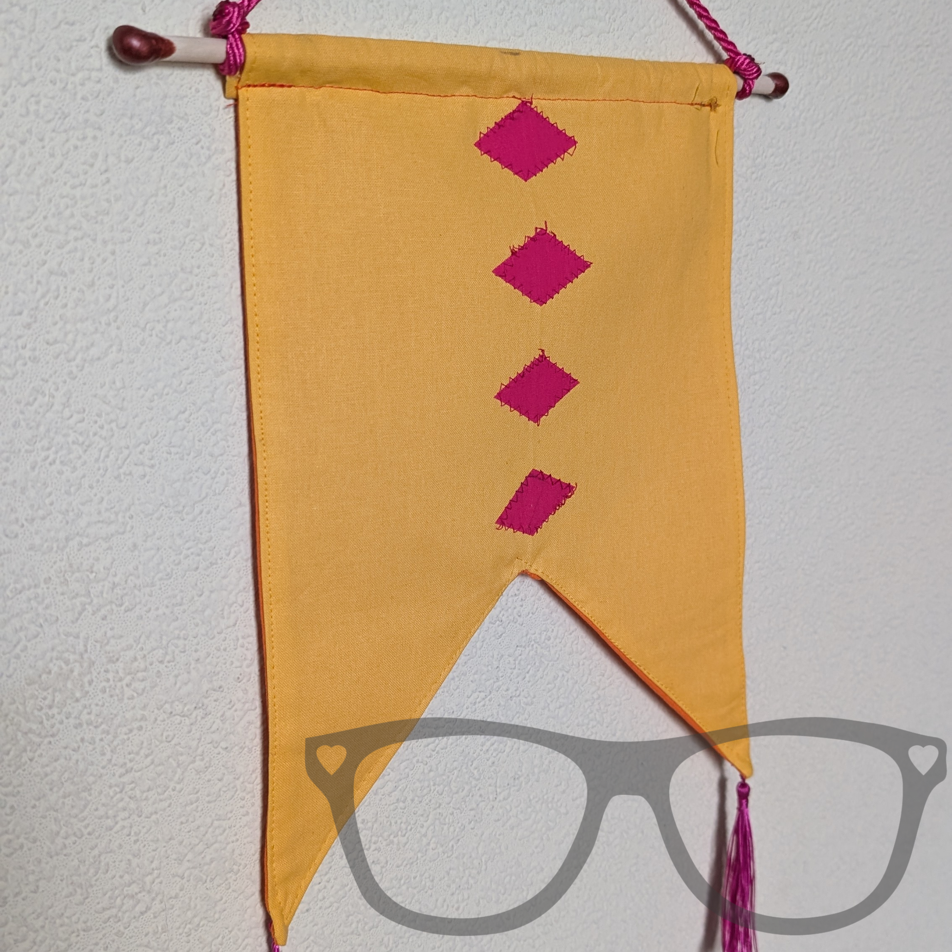 Yellow pennant style banner with 4 pink diamonds down the middle with two pink tassels. Hung on the wall using a cord and dowel dipped in wax.