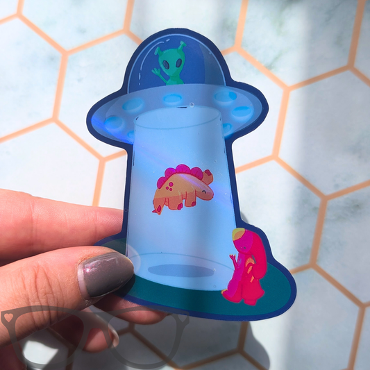 A hand holding a die-cut sticker or decal shaped like a UFO with cartoon aliens and a pink dinosaur on a light marble surface with orange hexagonal tile lines.