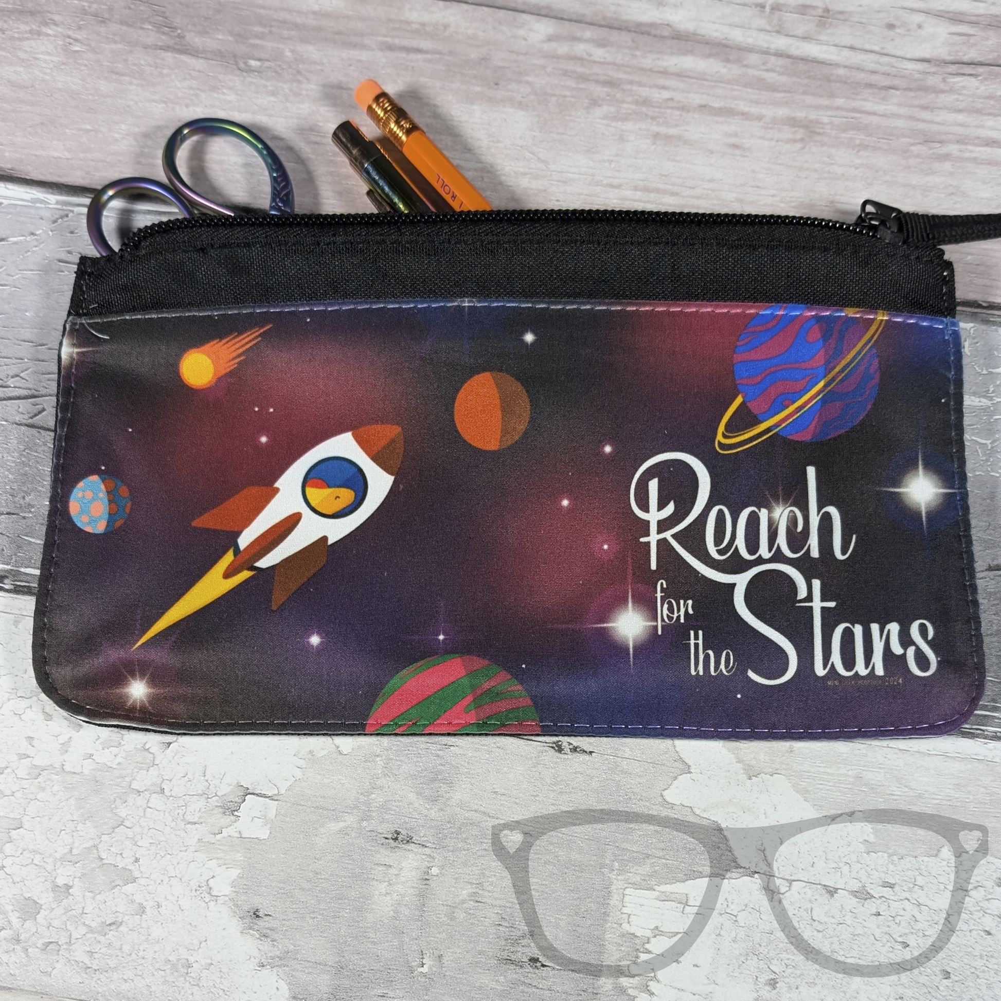 reach for the stars pencil case featuring an illustration of Derek the stegosaurus dinosaur going to space in a rocket shown with some stationery (not included)