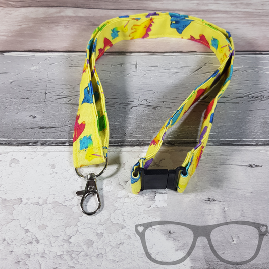 Dinosaur Lanyard, yellow fabric with colourful fun dinosaurs in a pattern. The lanyard features a lobster style clip for your keys or ID Card, and a quick release safety clip