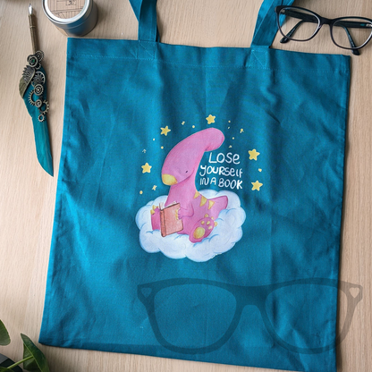 Flat lay of green cotton tote bag featuring Irene the dinosaur reading a book while sitting on a cloud.