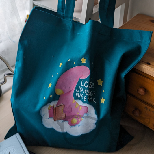 Green cotton tote bag with long handles featuring "Lose Yourself in a Book" design of Irene reading a book while sitting on a cloud.