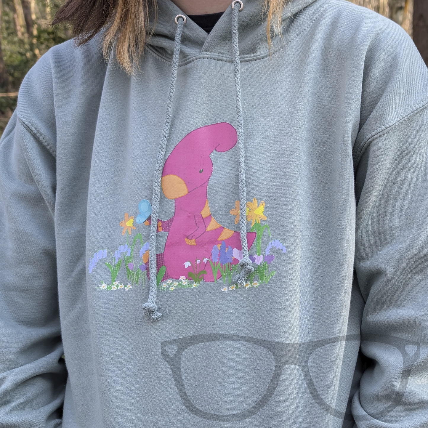 A soft dusty green hoodie featuring a digitally drawn design of Irene the Parasaurolophus sitting among spring flowers. This cosy dinosaur hoodie is perfect for geeks and dino fans of all ages