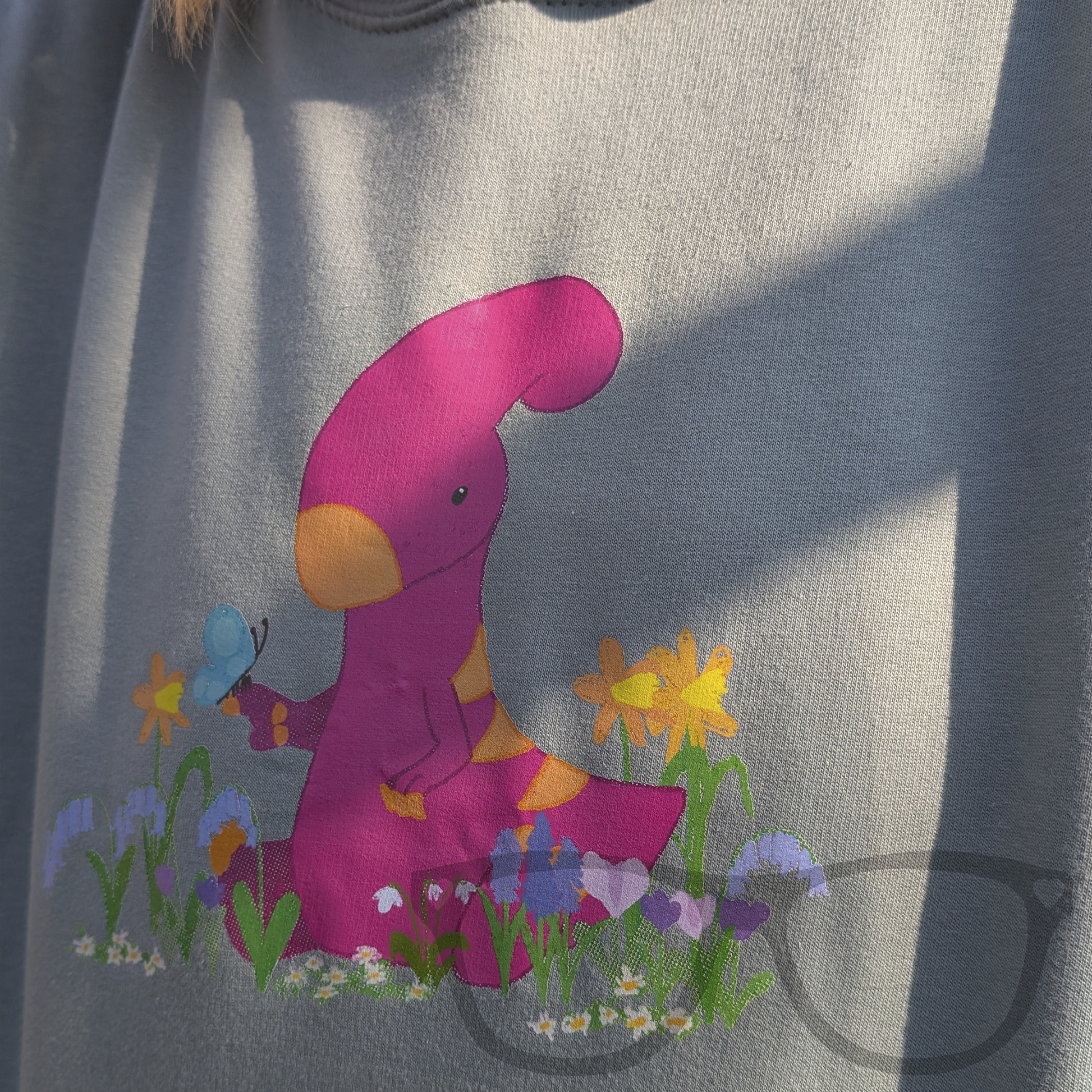 Detailed close-up of the Irene the Parasaurolophus spring design on a dusty green hoodie, showing the vibrant flowers and cute dinosaur illustration.