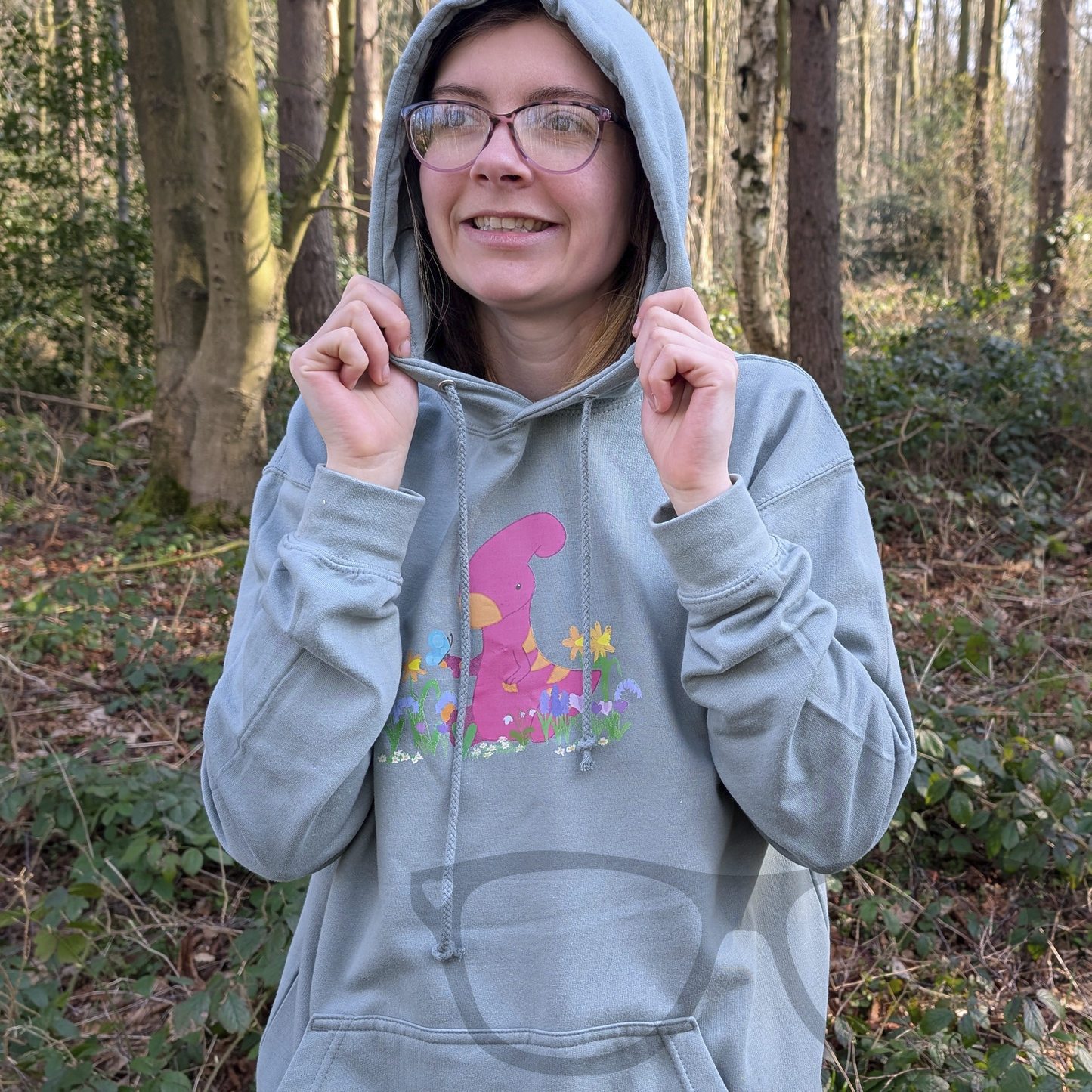 The dusty green dinosaur hoodie with the hood up, featuring Irene the Parasaurolophus in spring flowers. A perfect cosy and stylish look for any dino fan