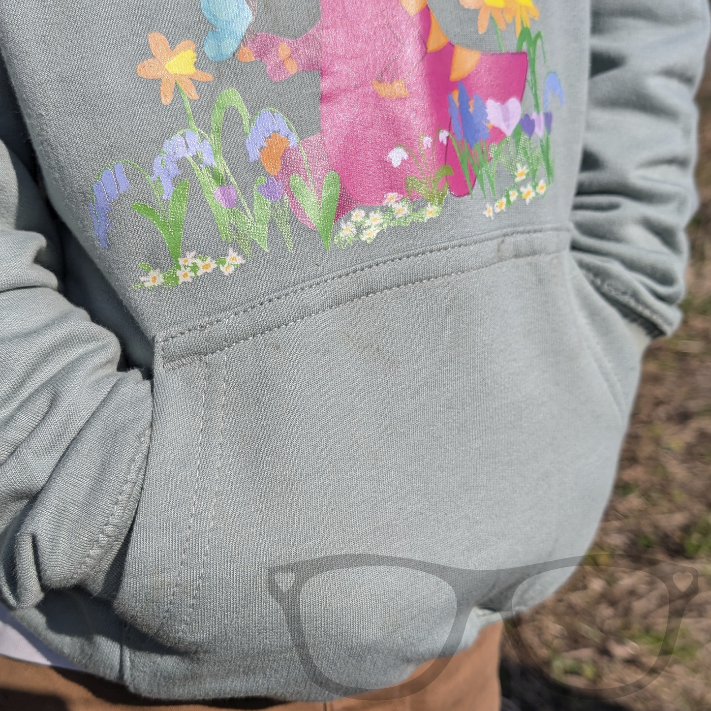 A child wearing the dusty green dinosaur hoodie with the kangaroo pocket on display. The hoodie features Irene the Parasaurolophus in spring flowers, offering comfort and style for young dino lovers.