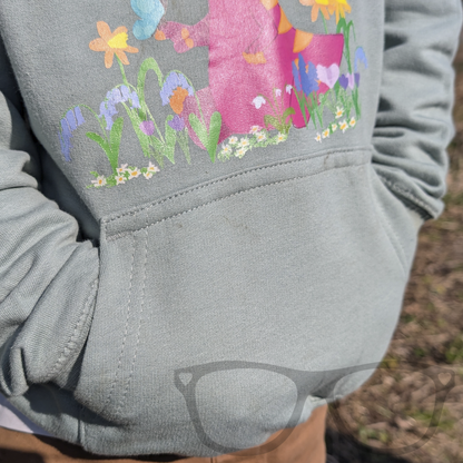 A child wearing the dusty green dinosaur hoodie with the kangaroo pocket on display. The hoodie features Irene the Parasaurolophus in spring flowers, offering comfort and style for young dino lovers.