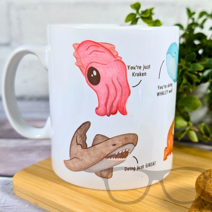 Motivational Marine Life mug showing cute illustrations of various marine animals with motivational phrases, this iamge shows a pink Cuttlefish saying "You're just kraken and a shark saying "Doing just GREAT"