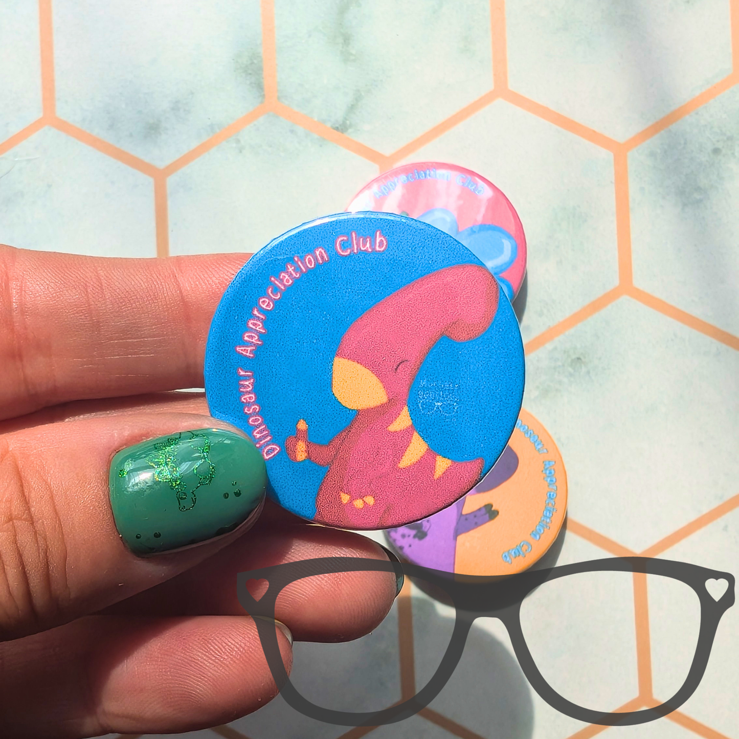 A hand with green glitter nail polish holds a round blue pinback button featuring a pink dinosaur and the text 'Dinosaur Appreciation Club'. Other colorful round buttons are stacked behind it on a light background with a hexagonal pattern, and a black eyeglasses silhouette is overlaid at the bottom.