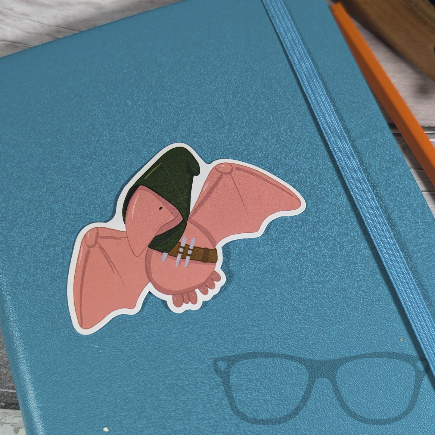 Pterry the Pterodactyl Rogue vinyl sticker on a blue notebook. A peach coloured stylised illustration of a Pterodactyl with a green hood and Rogue items