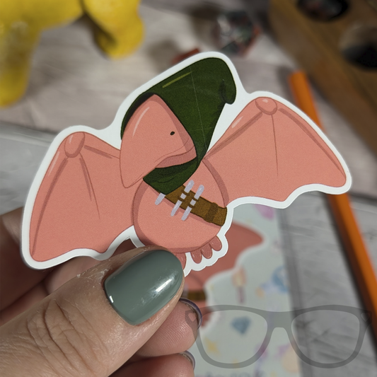 Pterry the rogue dinosaur and dnd vinyl die cut sticker  being held. A peach coloured stylised illustration of a Pterodactyl with a green hood and Rogue items