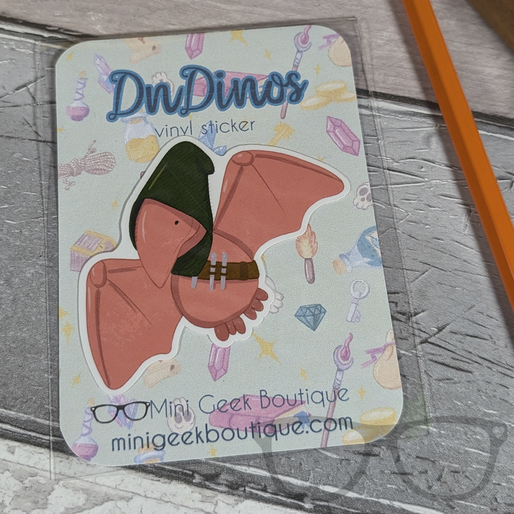 A dinosaur vinyl die cut sticker in the style of a peach coloured stylised illustration of a Pterodactyl with a green hood and Rogue items, in eco friendly packaging.
