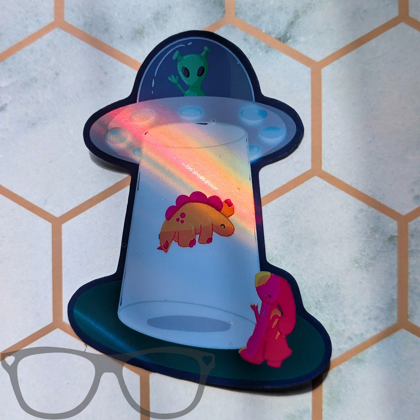 A novelty sticker or decal shaped like a UFO spaceship, featuring a green alien in the cockpit, a pink and orange dinosaur inside, and a small pink Parasaurolophus outside, displayed on a light grey surface with a hexagonal pattern.