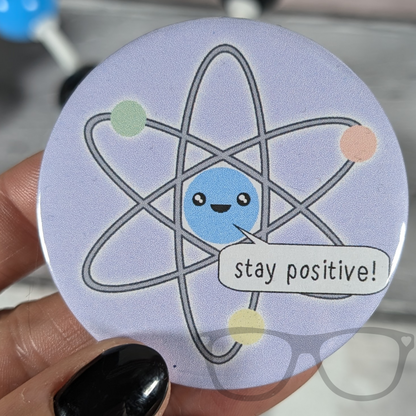 Stay positive atomic science badge, purple badge with cute kawaii atom saying "Stay Postitive"