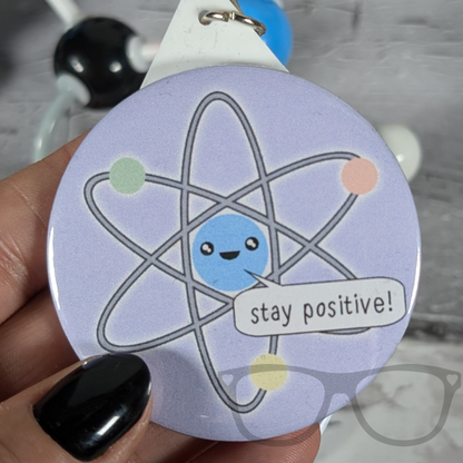 Stay positive atomic science keyring, purple badge with cute kawaii atom saying "Stay Postitive"