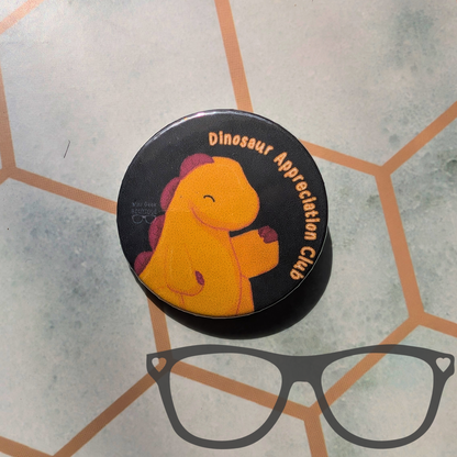 A round black pinback button or sticker featuring a cartoon yellow dinosaur with purple spikes and the text 'Dinosaur Appreciation Club,' placed on a light grey patterned surface. A grey glasses outline graphic is overlaid at the bottom.