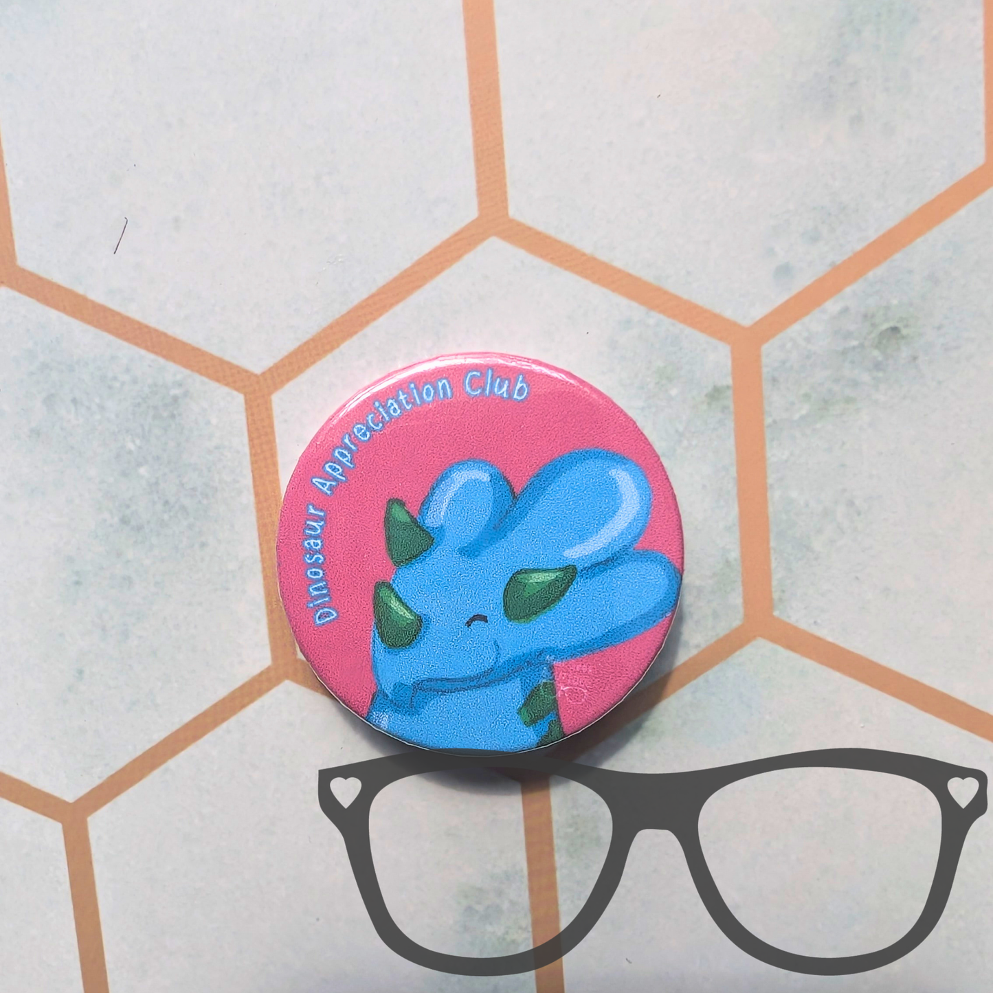 A round pin badge featuring a blue cartoon dinosaur with green horns on a pink background, with the text 'Dinosaur Appreciation Club' around the edge. The badge is placed on a light grey surface with a hexagonal pattern and a black eyeglasses graphic below it.