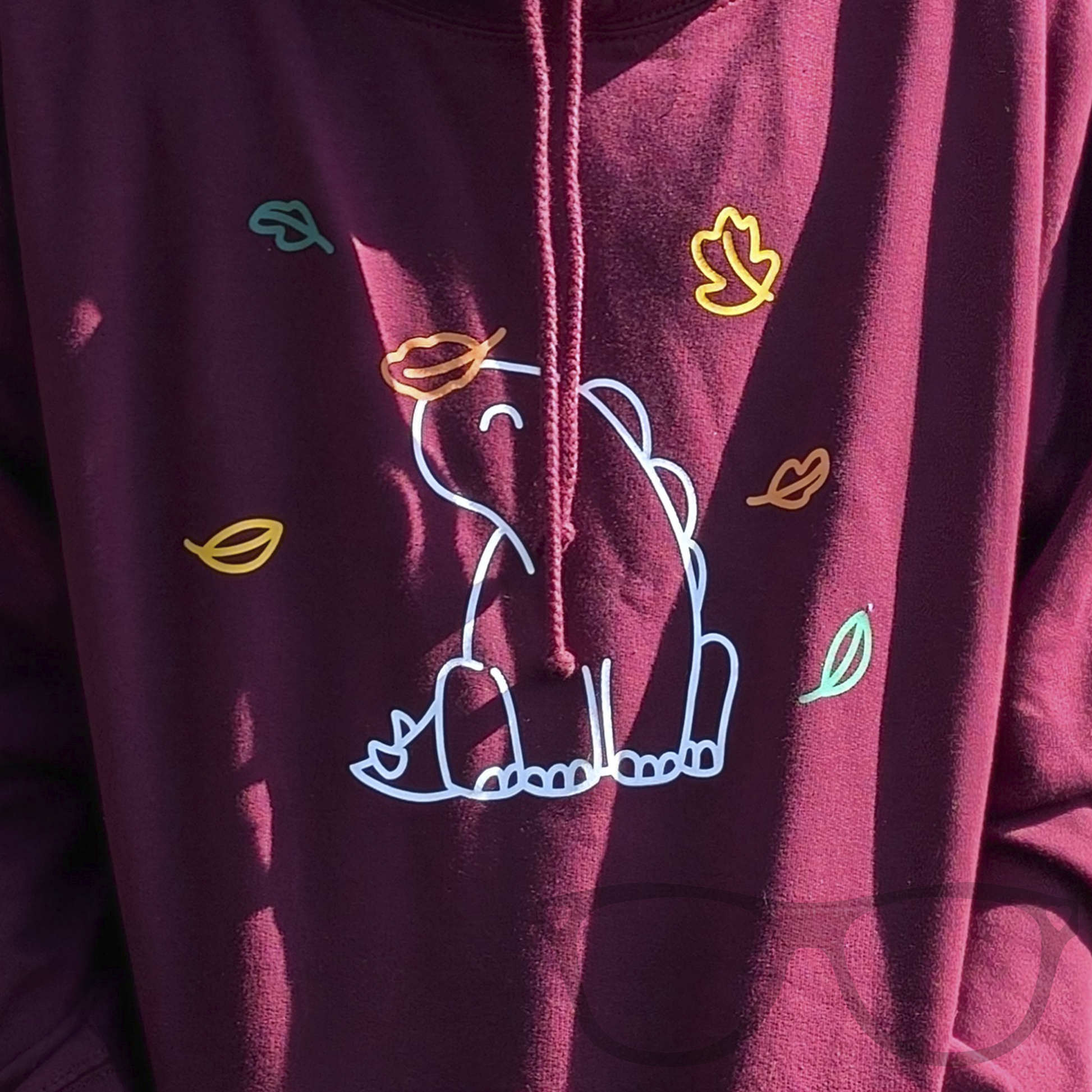 Close up detail of Burgundy Hoodie with kangaroo pocket for Autumn featuring Derek the Stegosaurus sitting amongst the falling autumn leaves