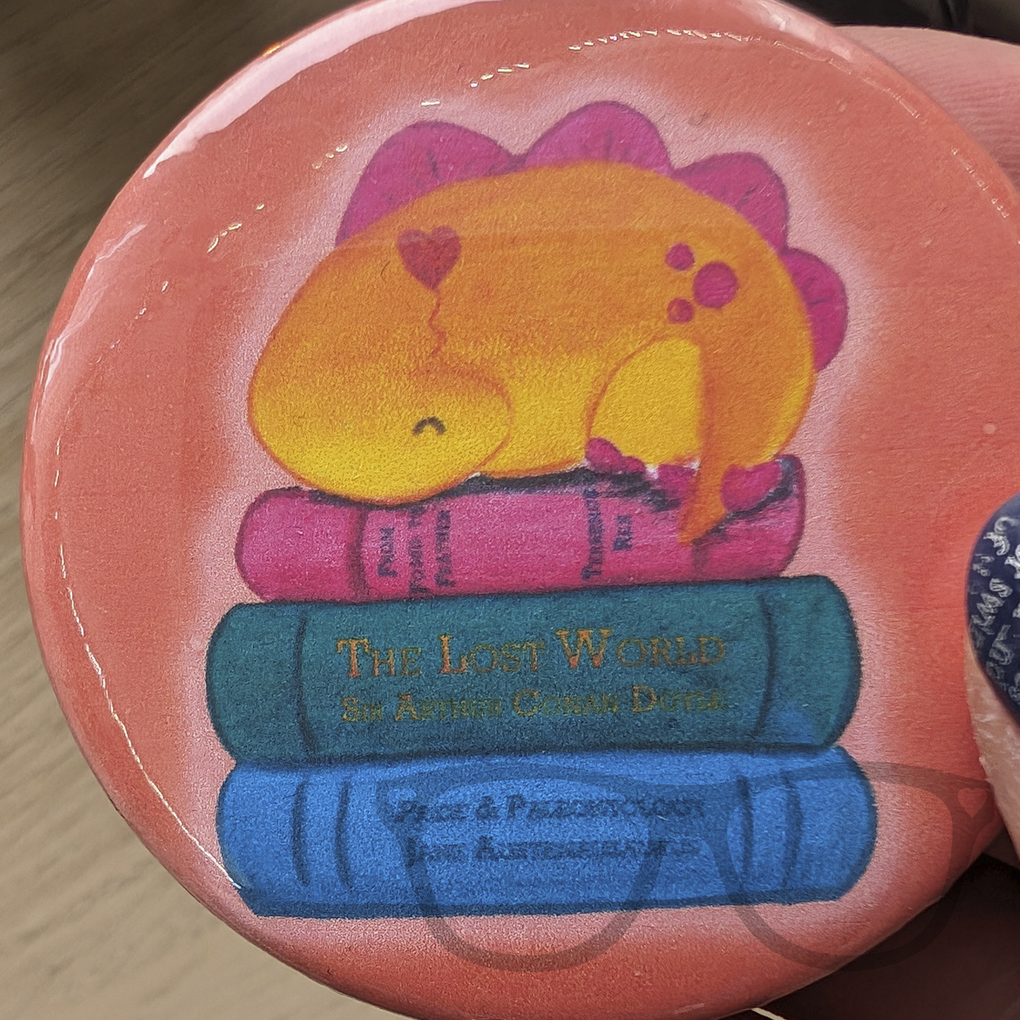 An orange circular 58mm button badge featuring derek the dinosaur napping on a pile of books