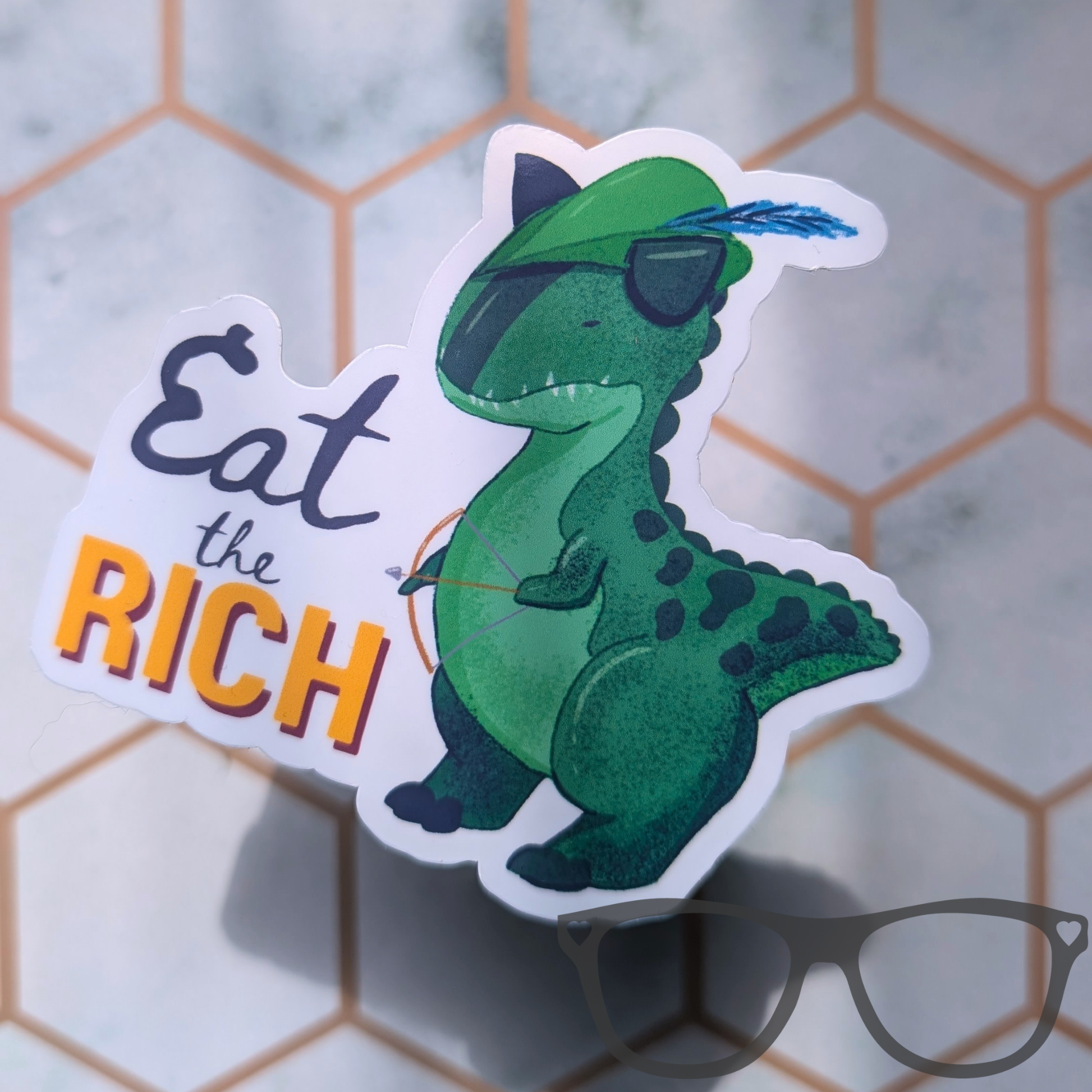 A die-cut sticker featuring a green illustrated Carnotaurus a Robin Hood  and a blue feather in its green hat, next to the text 'Eat the RICH' in grey and orange lettering. 