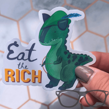 A die-cut sticker featuring a green cartoon dinosaur wearing a Robin Hood style hat with a blue feather, with the text 'Eat the RICH' in dark blue and orange. The sticker is held by a hand against a blurred light grey background with a hexagonal pattern.
