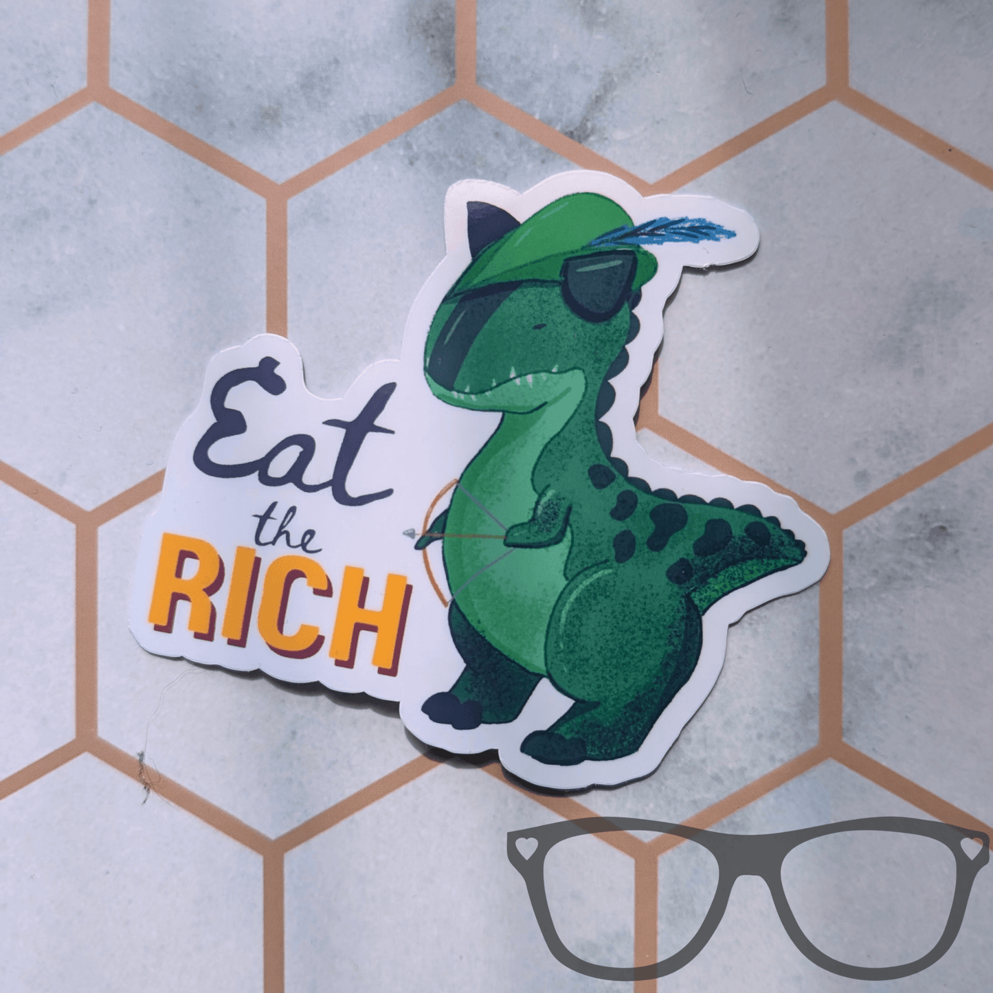 A die-cut sticker featuring a green cartoon dinosaur wearing a Robin Hood feathered hat, holding a bow and arrow next to the text 'Eat the RICH' in black and orange, placed on a light grey surface with a hexagonal pattern.