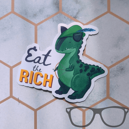 A die-cut sticker featuring a green cartoon dinosaur wearing a Robin Hood feathered hat, holding a bow and arrow next to the text 'Eat the RICH' in black and orange, placed on a light grey surface with a hexagonal pattern.