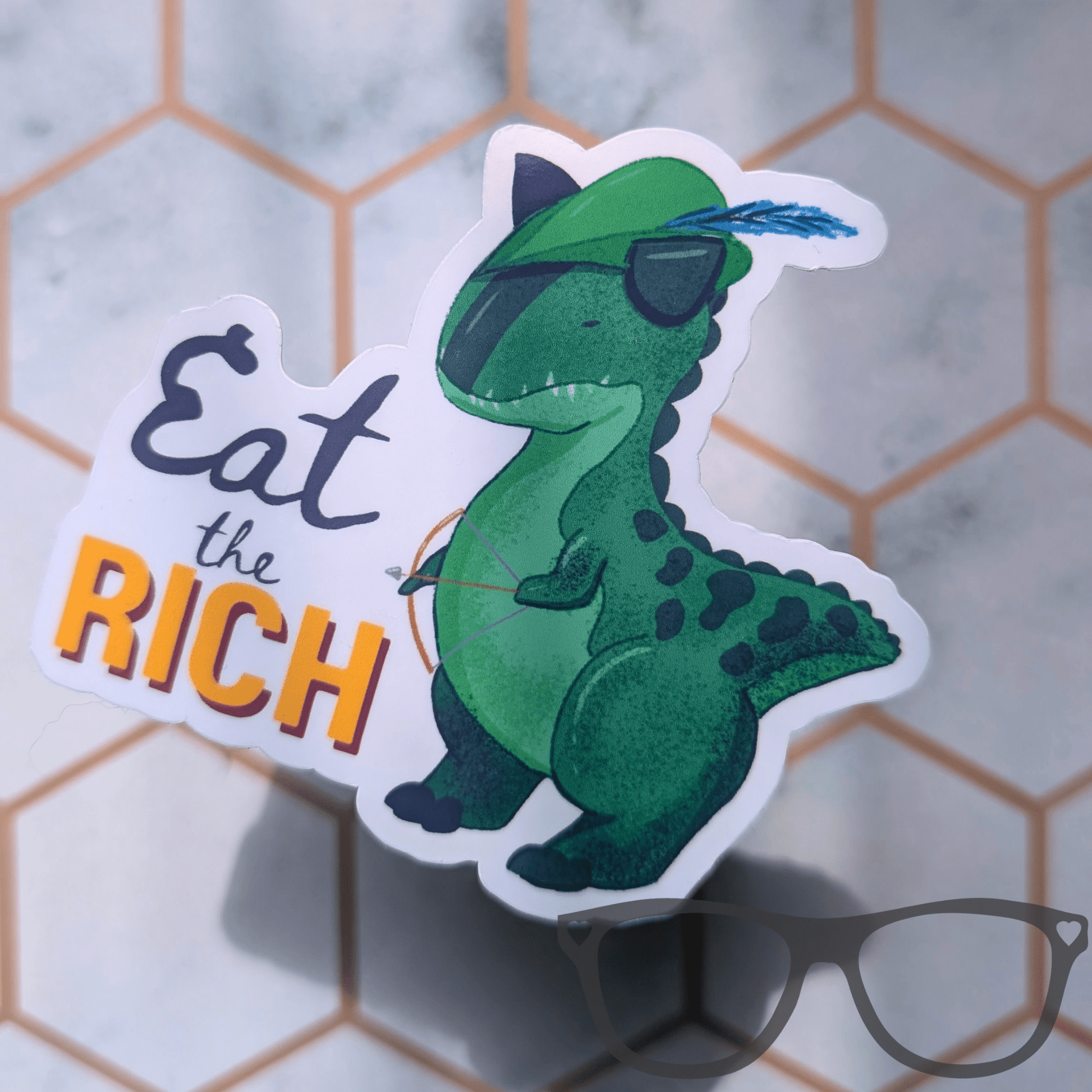 A die-cut sticker featuring a green cartoon T-rex wearing a Robin Hood style pointy hat with a blue feather, and holding a bow and arrow next to the text 'Eat the RICH' in black and orange lettering. The sticker is displayed on a light marble surface with a hexagonal grid pattern and a pair of glasses in the foreground