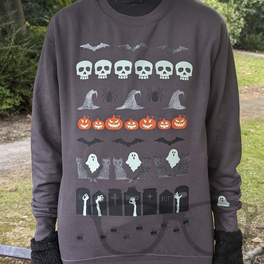Grey Halloween sweater featuring a variety of spooky graphics including skulls, bats, witches' hats, spiders, and pumpkins with glow-in-the-dark eyes, styled like a traditional ugly sweater.