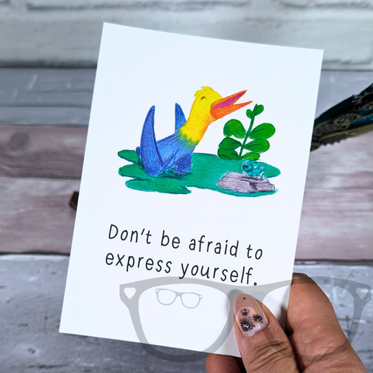 Motivational A6 postcard with Holly the quetzalcoatlus and the phrase "Don't be afraid to express yourself" on a glossy surface.
