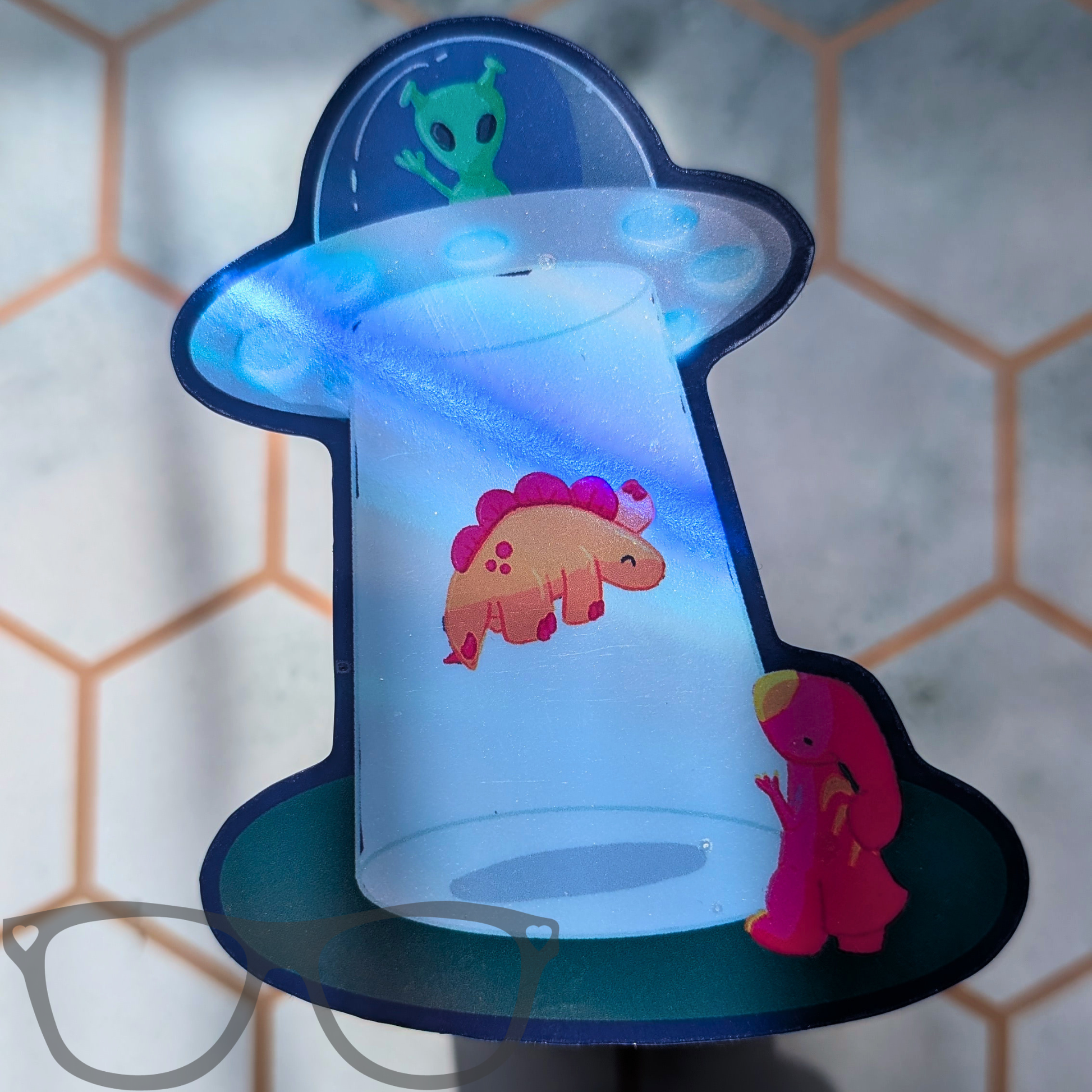 Holographic die cut sticker The design features a green alien waving from the dome, a pinkish-orange Stegosaurus dinosaur printed on the side, and a small pink dinosaur figure sitting next to the base.