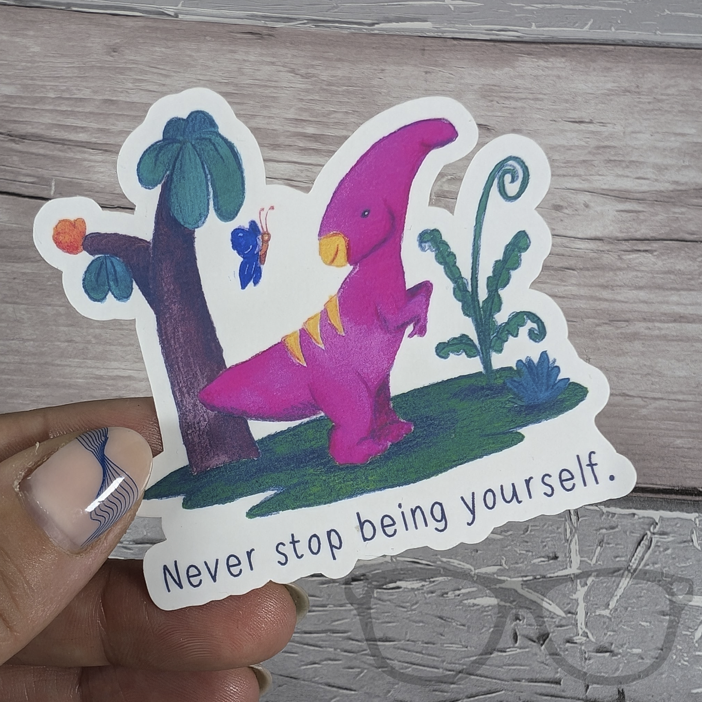 Irene the Parasaurolophus "Never Stop Being Yourself" sticker being held, a water-resistant motivational sticker perfect for laptops, water bottles, and more.
