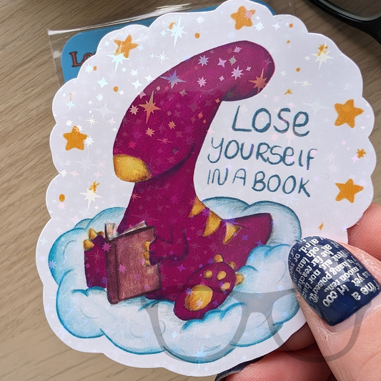 Sparkly sticker featuring a pink Irene the Parasaurolophus reading a book on a cloud, the text reads "Lose yourself in a book" the overlay of the sticker features stars and sparkles