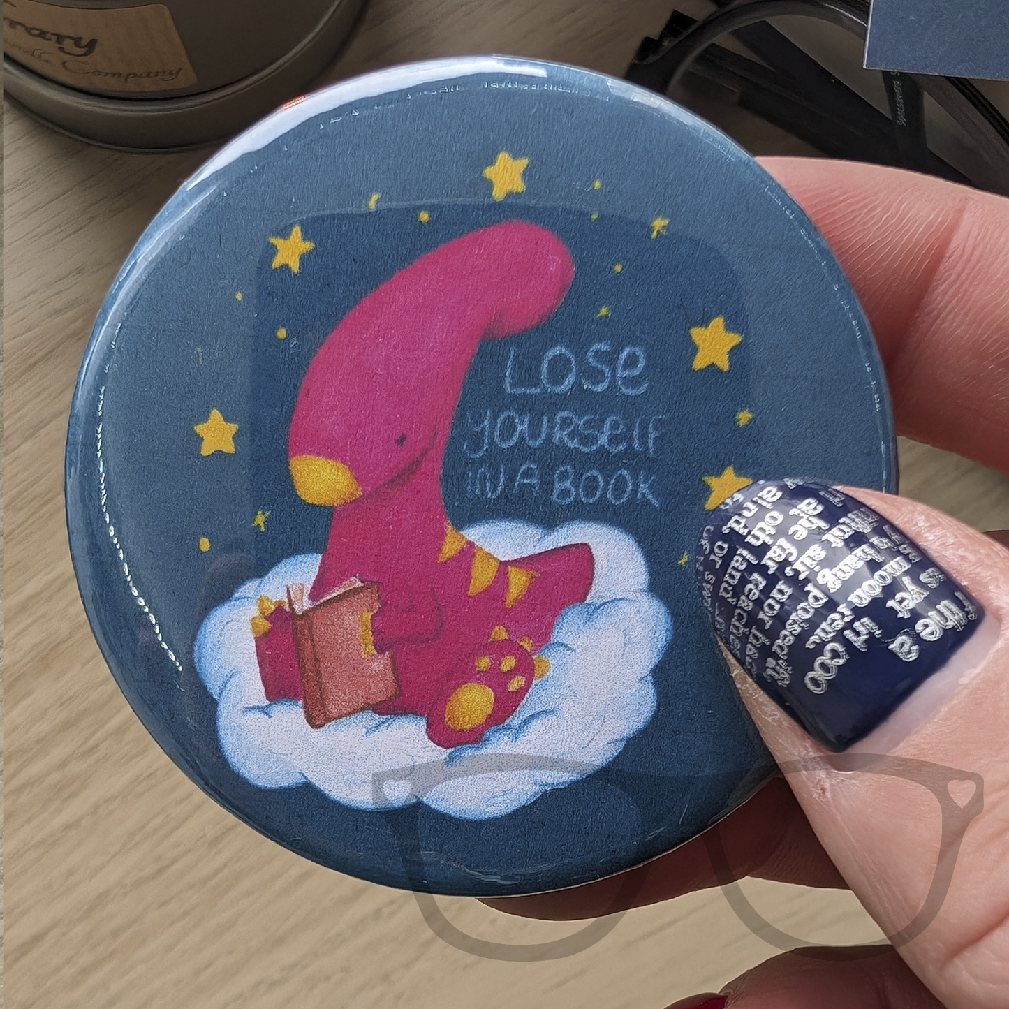 a 58mm circular button badge featuring Irene the Dinosaur on a cloud reading a cook. The text reads "Lose yourself in a book"