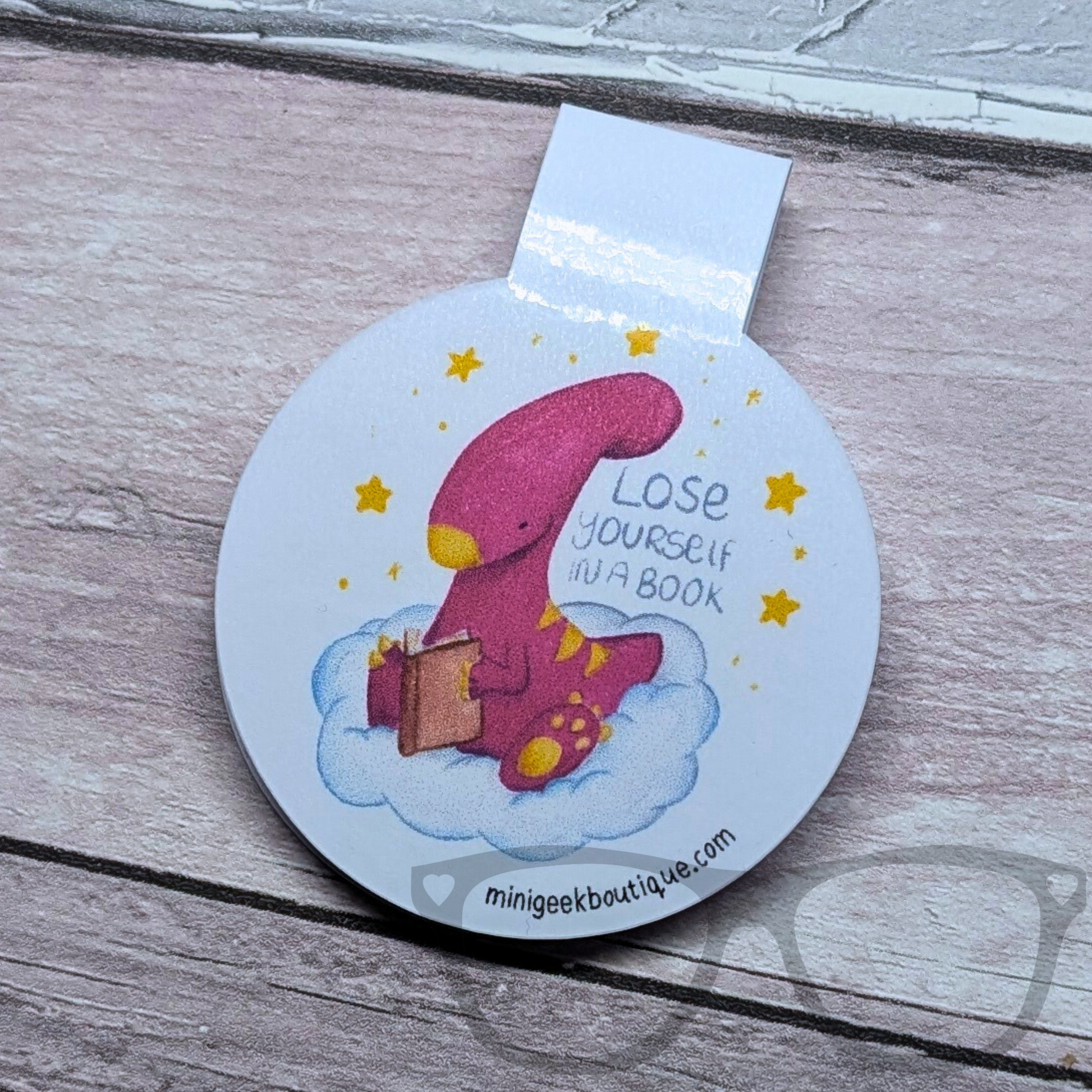 Magnetic bookmark featuring Irene the Parasaurolophus reading a book on a cloud. Gloss laminated and designed to mark your place without damaging pages.