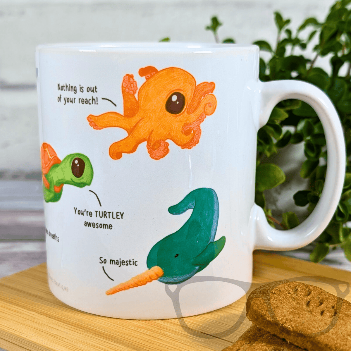 Mug with cartoon octopus, turtle, and narwhal design on a wooden surface with cookies.