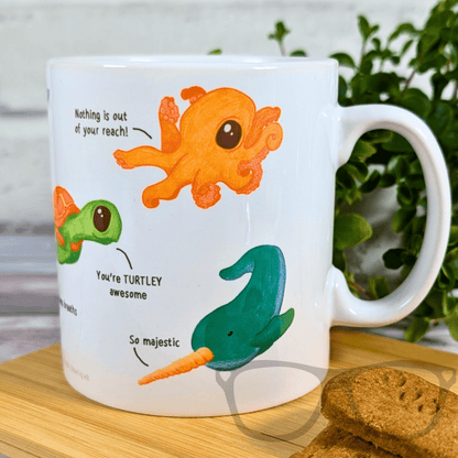 Mug with cartoon octopus, turtle, and narwhal design on a wooden surface with cookies.
