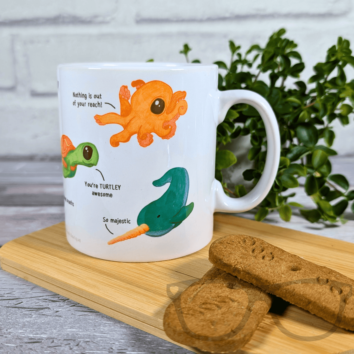 White mug with cartoon octopus and turtle design on a wooden board with cookies and a plant in the background