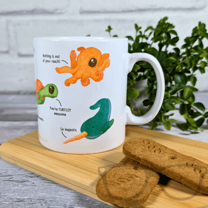 White mug with cartoon octopus and turtle design on a wooden board with cookies and a plant in the background