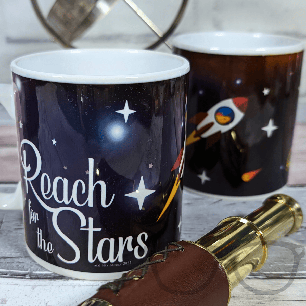 Two mugs with space-themed designs and a telescope on a wooden surface.