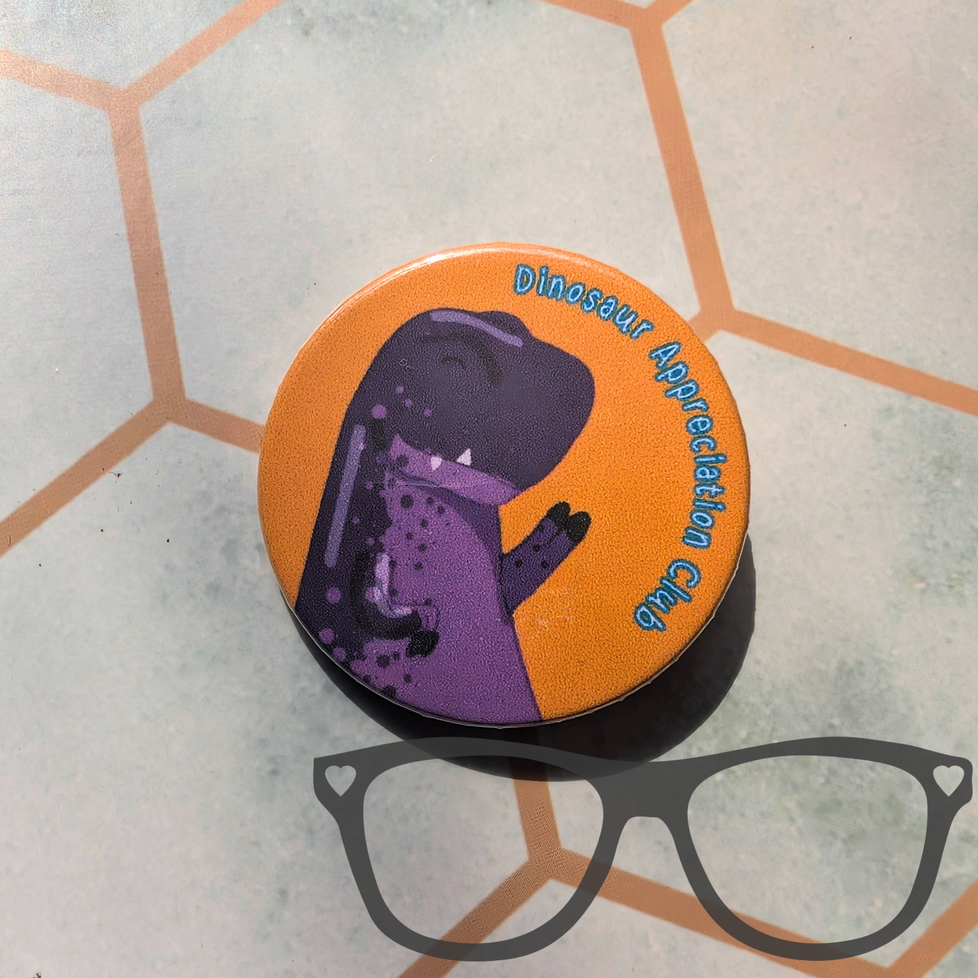 A round pin badge featuring a purple dinosaur on an orange background with the text 'Dinosaur Appreciation Club', placed on a light grey surface with a hexagonal pattern and a black-framed glasses graphic overlay.