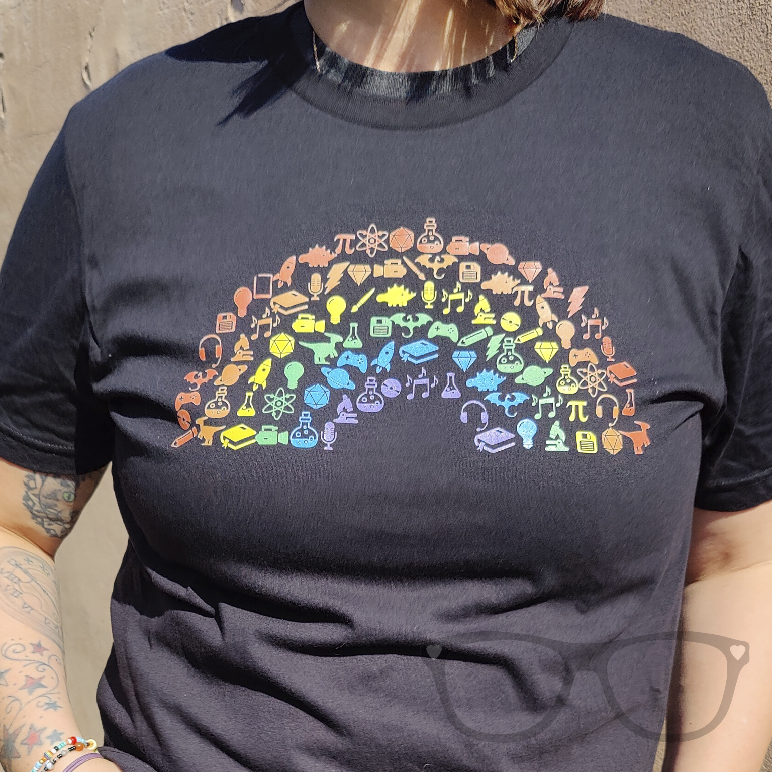 Adult model wearing the Geek and Proud T-shirt featuring a rainbow design with geeky icons like game controllers, dice, and planets.
