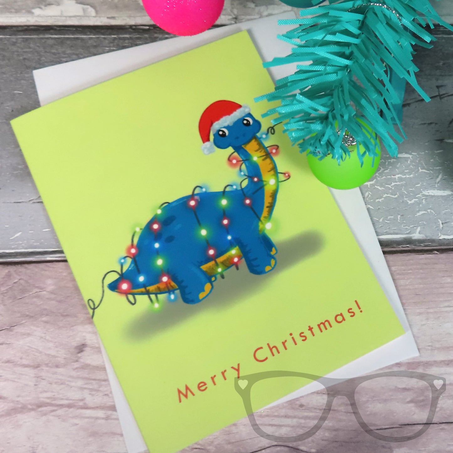 Dinosaur Christmas greeting card, features Brenda the Brachiosaurus tangled up in some fairy lights. Car dis shown flat on the surface with an envelope underneath