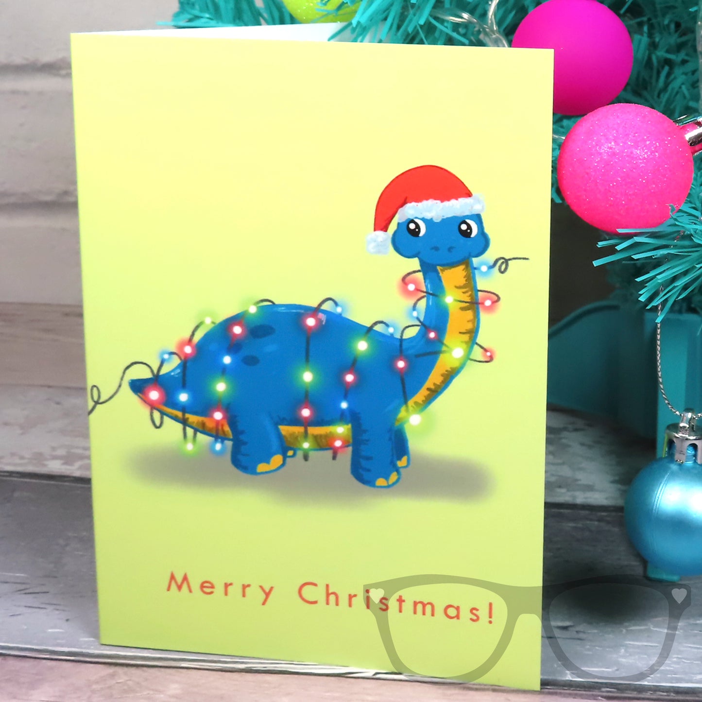 Christmas greetings card with Brenda the dinosaur on, card is standing against a festive tree